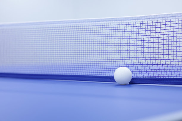 一颗白色乒乓球放在乒乓球桌上，后面有一个网。背景模糊的白色乒乓球。图片下载