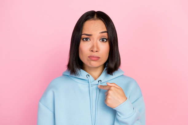 年轻女孩怀疑地指着自己，扬起眉毛，身穿蓝色时装，粉色背景照片图片下载