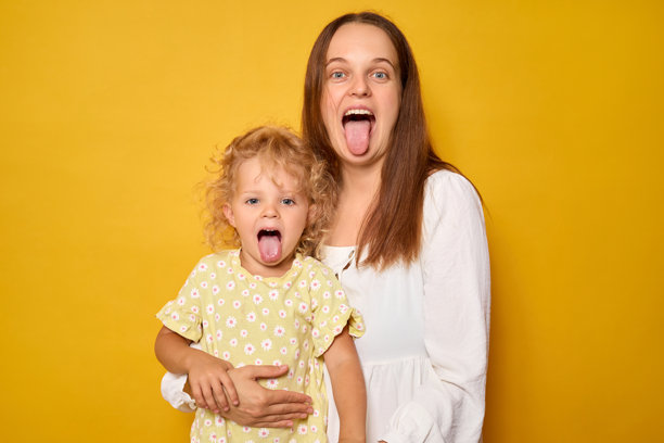 兴奋的女人和孩子在黄色背景下分享亲密而有趣的时刻，家庭成员表现出顽皮的行为，吐出舌头并做出搞怪的表情。图片下载