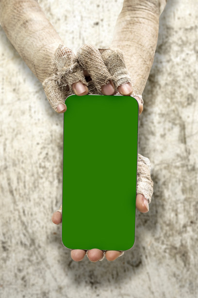 木乃伊的手缠着脏绷带，拿着带绿色屏幕的手机，背景是粗糙的墙壁图片下载