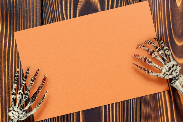 人类骨骼手在木质背景上拿着橙色纸片，空白橙色纸片可用于复制空间，图片具有令人毛骨悚然和不安的气氛，恐怖图片下载