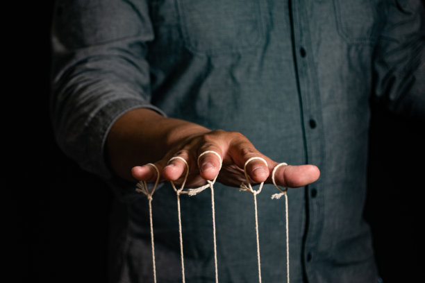 特写镜头：男人用手指操控木偶。男人的手通过与线相连的手指控制木偶，背景为黑色，体现操控的概念。图片下载