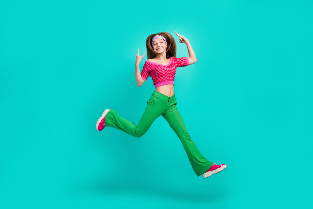 穿着粉色短款上衣和喇叭裤的漂亮酷女孩在青绿色背景下跳跃，做出摇滚手势的全身照片图片下载