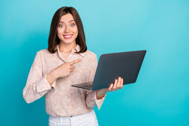 可爱的年轻女性在青色背景下，穿着时尚休闲服装，指着笔记本电脑进行远程工作的人像照片。图片下载