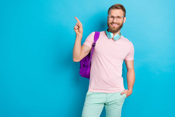 年轻男子的照片肖像，佩戴眼镜，背着背包，指向空白空间，穿着时尚的粉色服装，背景为青色。图片下载