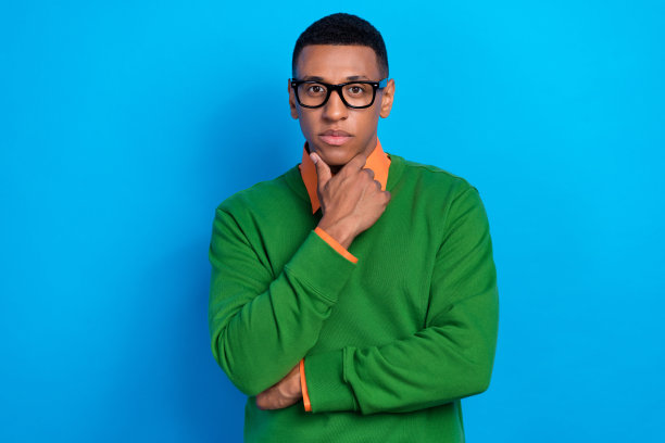 穿着橙色衬衫和绿色毛衣的自信聪明男士，手托下巴，背景为蓝色。图片下载