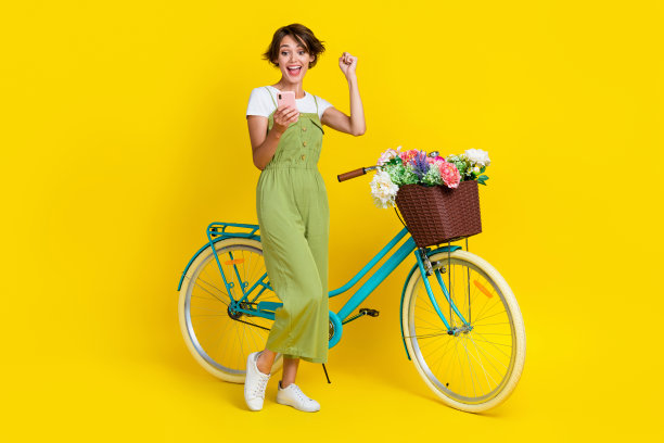 全身照，年轻可爱的女孩骑自行车，手持设备像是在评论，穿着时尚的卡其色服装，背景为黄色。图片下载