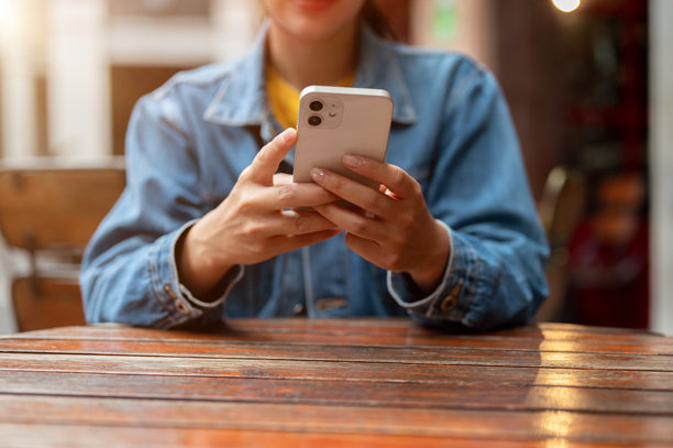 一张裁剪过的图片，展示了一位穿着牛仔夹克的女性在户外桌子旁使用智能手机。图片下载