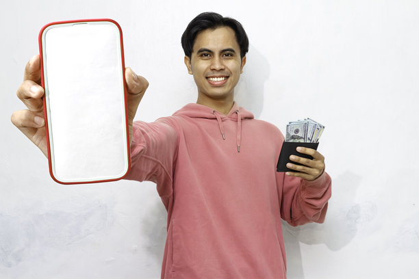 穿着粉色连帽衫的亚洲男子兴奋地微笑，手里拿着装满现金的钱包，盯着一部空白屏幕的智能手机，背景为白色。图片下载