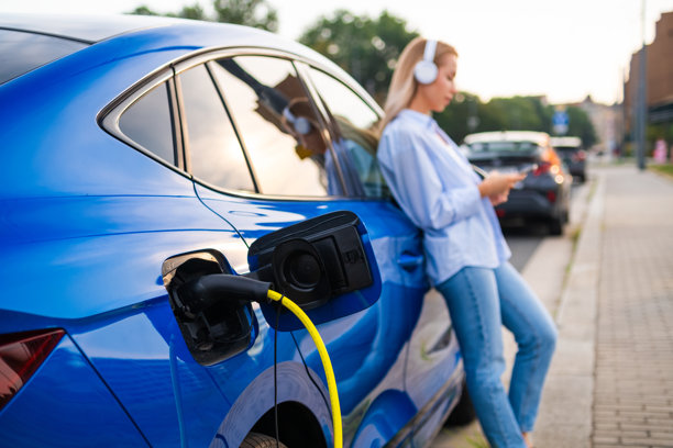 女人戴着耳机站在电动汽车旁，在路边充电图片下载