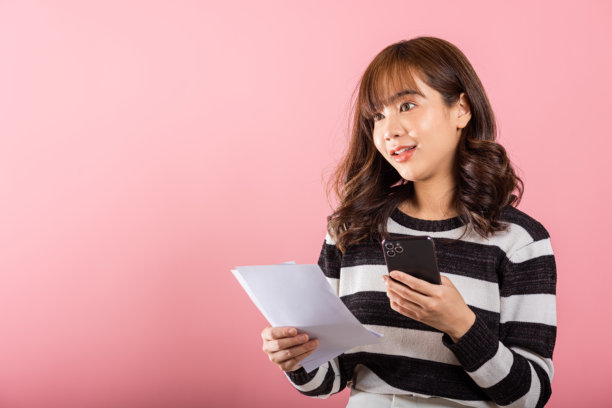 快乐的亚洲年轻女性手持手机，查看家庭预算账单和在线支付发票，享受便捷的网上银行服务。图片下载