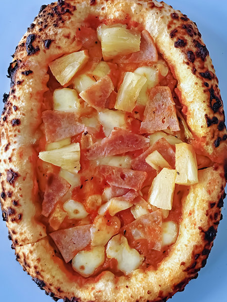 整张图片展示了一整块新鲜出炉的椭圆形夏威夷披萨，酸面团底，顶部覆盖番茄酱、马苏里拉奶酪、火腿块和菠萝块，背景为蓝色，俯视角度。图片下载