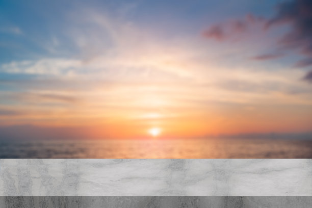 日落时分的空白白色大理石桌子，背景是模糊的海滩和海洋散景，前景有绿色叶子，适合用作社交媒体广告的产品展示或内容拼贴模板。图片下载