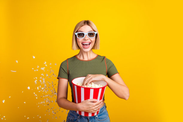戴着3D眼镜、穿着卡其色T恤的短发女孩，惊讶地吃着爆米花观看首映，背景为黄色。图片下载
