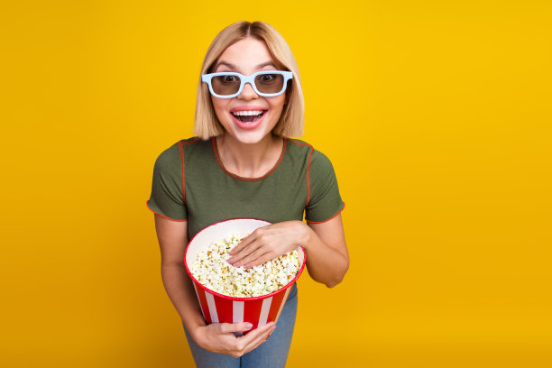 一个留着波波头的女孩，穿着卡其色T恤，戴着3D眼镜，吃着爆米花，观看电影，背景为黄色。图片下载