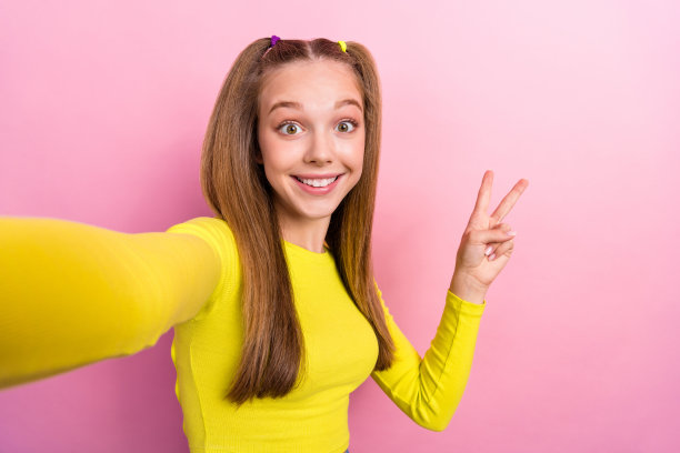 可爱的快乐女孩扎着马尾，穿着黄色长袖，录制视频并做出V字手势，背景为黄色。图片下载