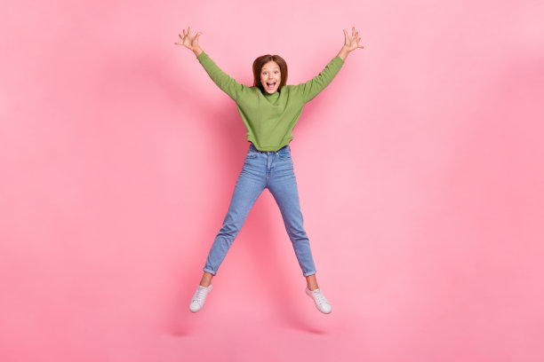 穿着绿色卫衣的搞笑兴奋女学生在粉色背景下高高跳起，展现出明星般的姿态的全身照片图片下载