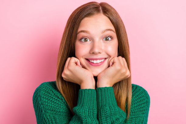 特写照片：年轻梦幻、快乐的漂亮女孩，棕色头发，双手托腮，面带积极的灿烂微笑，孤立在粉色背景上。图片下载