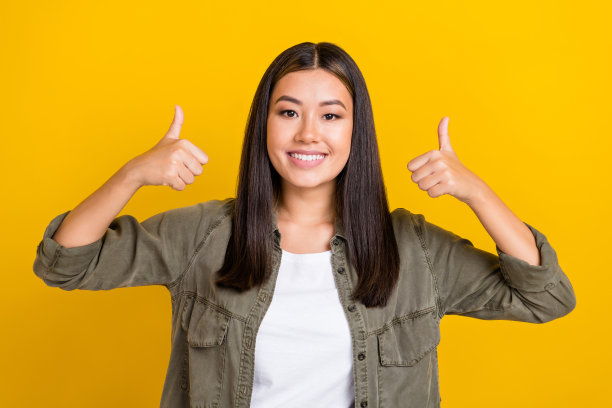 满意、愉快的女孩肖像，长发，穿着卡其色衬衫，竖起大拇指，背景为黄色。图片下载