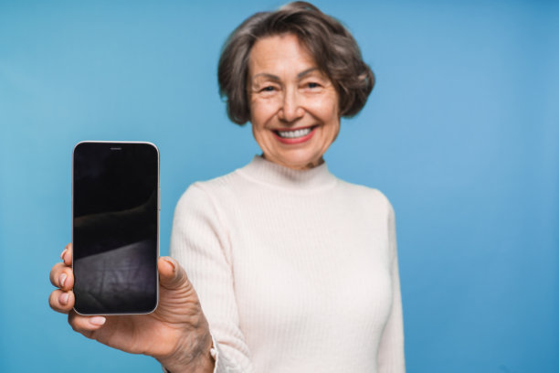 快乐的白人老年女性使用手机，展示空白的手机屏幕，背景为单色。图片下载