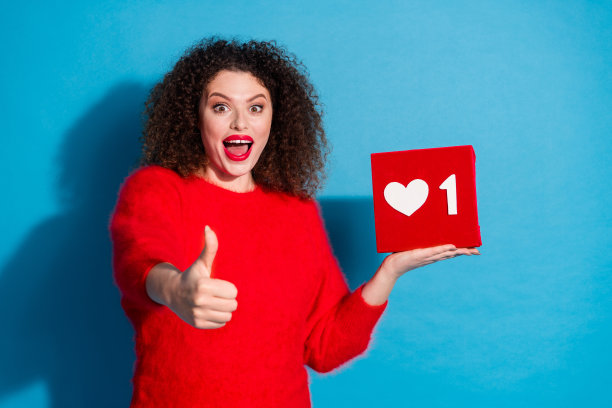 穿着时尚红色衣服的迷人年轻女性，手持社交媒体图标并竖起大拇指，背景为蓝色。图片下载