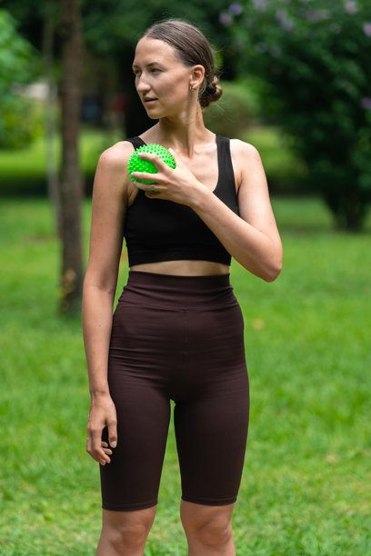 年轻女性穿着运动服在公园里用带刺的绿色按摩球按摩肩膀，促进放松和缓解肌肉紧张。竖版照片图片下载