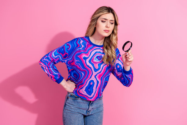 一位有趣、好奇、严肃的女士的肖像照片，染发，穿着彩色衬衫，手持放大镜观察物体，背景为粉色。图片下载