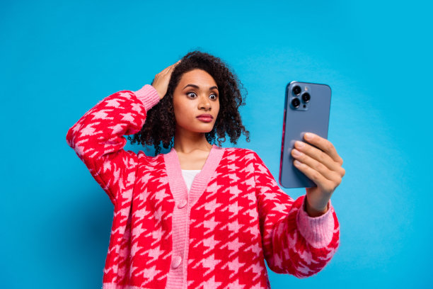 一位美丽的美国女士在蓝色背景下，困惑地用手触摸头部，凝视着智能手机镜头的肖像照片。图片下载