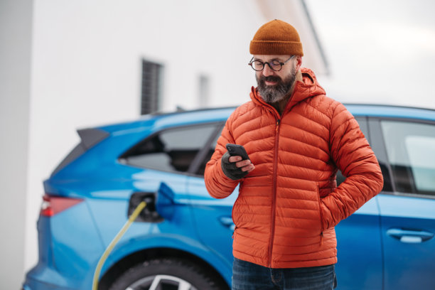 在寒冷的雪天，男子正在为电动车充电，使用电动车充电应用程序，查看电池电量和能耗。冬季充电和驾驶电动车。图片下载