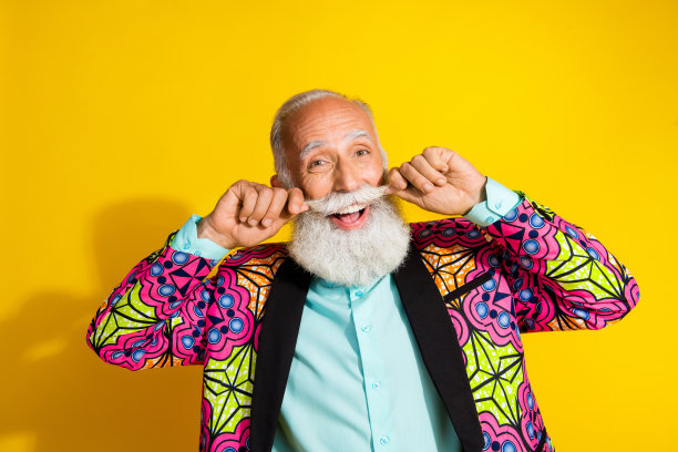 穿着时尚、色彩鲜艳的粉色华丽服装的迷人退休男士，留着胡须，站在黄色背景前的理发店照片图片下载