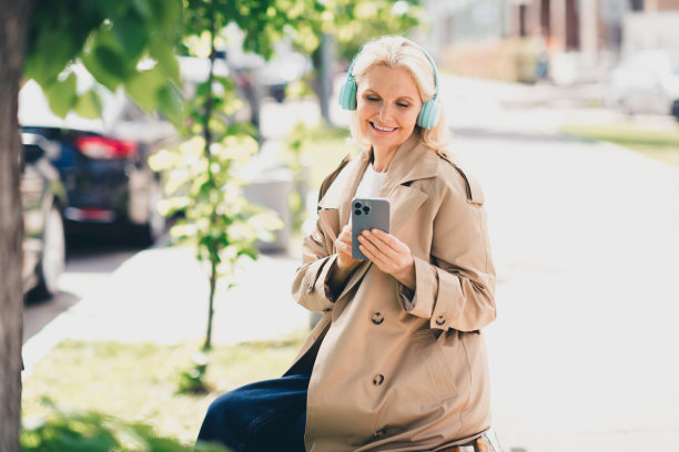 快乐的退休女性在春季周末户外公园街道上，穿着米色外套，戴着耳机听音乐。图片下载