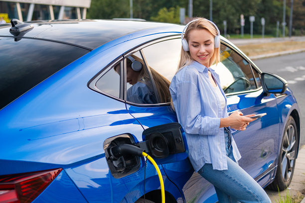 年轻女子戴着耳机听音乐，倚靠在充电的电动汽车旁图片下载