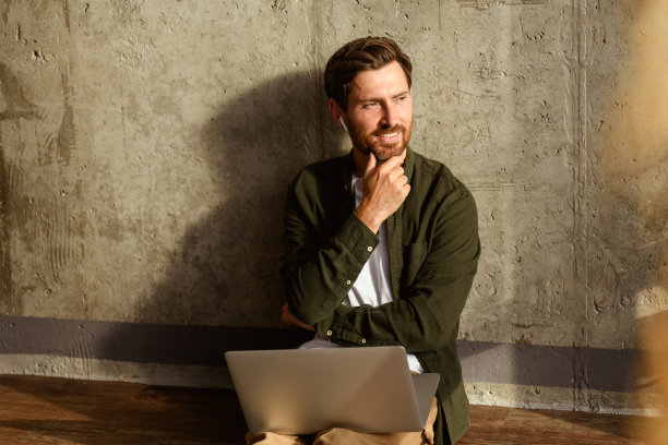 微笑的室内设计师戴着耳机，手持笔记本电脑，站在公寓的混凝土墙旁。图片下载