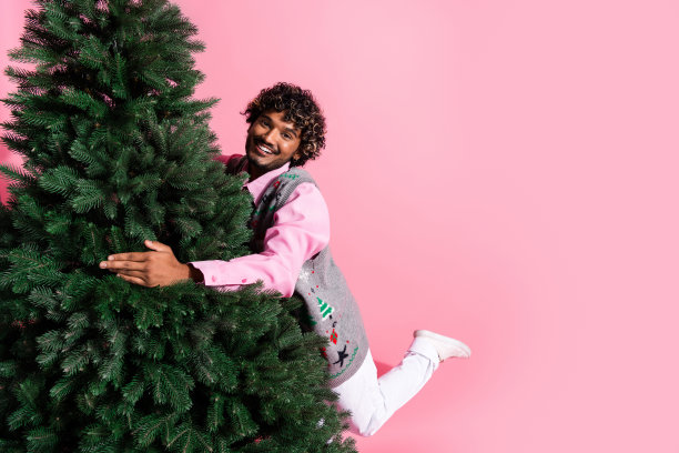 穿着花哨背心的酷炫男士在粉色背景下的空旷空间里购买圣诞树的照片图片下载