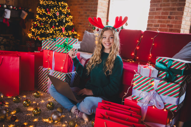 穿着绿色圣诞服装的少女在家中派对装饰的客厅里，手持电子商务笔记本和信用卡，拍摄的全身照片。图片下载