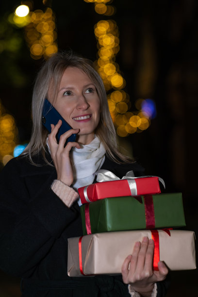 美丽年轻快乐的金发女性在夜晚的城市中走动，一边打电话一边手持礼品盒。节日、圣诞节的概念。竖版照片图片下载