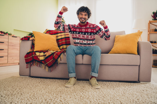 年轻男子坐在沙发上，举起拳头，穿着圣诞毛衣，庆祝节日的室内公寓全景照片图片下载
