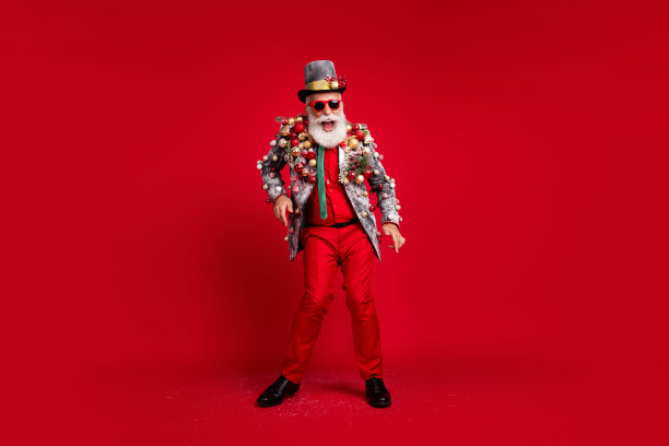 全身照片的圣诞老人，搞笑的男人穿着时尚的服装，跳舞，身边有圣诞树和装饰球，戴着太阳镜，背景为红色。图片下载