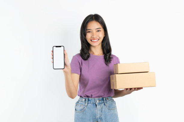 微笑的亚洲女性手持智能手机，旁边有纸箱，背景为白色，在线配送概念。图片下载