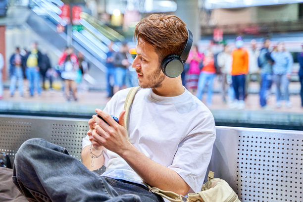 年轻的拉丁裔男子在等火车，戴着耳机看着智能手机。图片下载