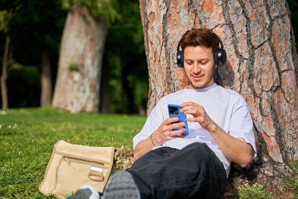 一位拉丁裔男子坐在公园的草地上，靠着一棵树。他放松身心，使用智能手机来摆脱日常压力。图片下载