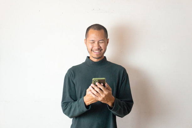 年轻的亚洲男子穿着冬季服装或毛衣，笑着拿着智能手机，背景为纯白色。图片下载