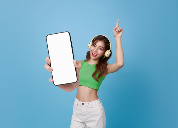 年轻快乐的亚洲女性戴着耳机，听着音乐，展示手机模型，背景为蓝色。图片下载