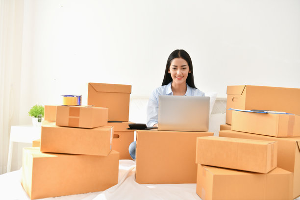 微笑的年轻女性在床上使用笔记本电脑，身边有纸箱，靠在家里的墙上。图片下载