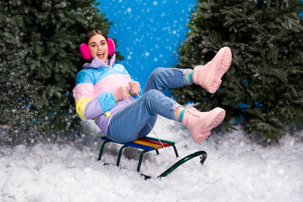 兴奋的时尚女士穿着条纹外套在室内蓝色背景下滑雪橇，周围是圣诞树。图片下载