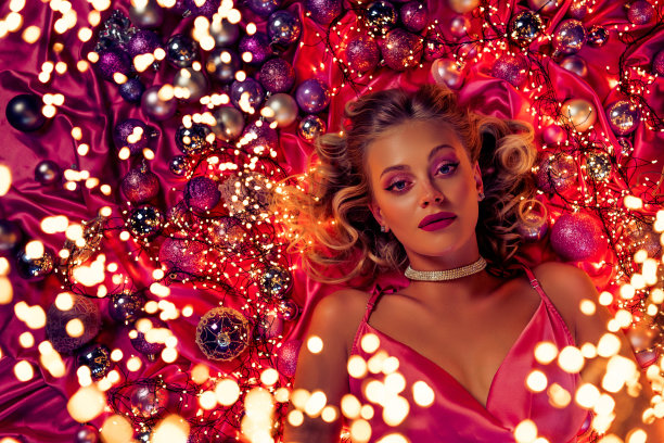 俯视照片中迷人的女士正在准备圣诞节活动，躺在粉色的树球和仙女灯光旁。图片下载