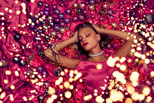 俯视照片中，一位迷人的派对女士躺在粉色丝滑的质地上，周围装饰着圣诞树的闪亮球饰。图片下载