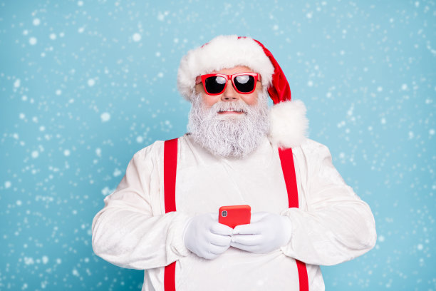 特写照片：搞笑的胖白发圣诞老人使用手机发送短信祝贺，穿着红色吊带工装，戴着太阳镜，背景为蓝色。图片下载