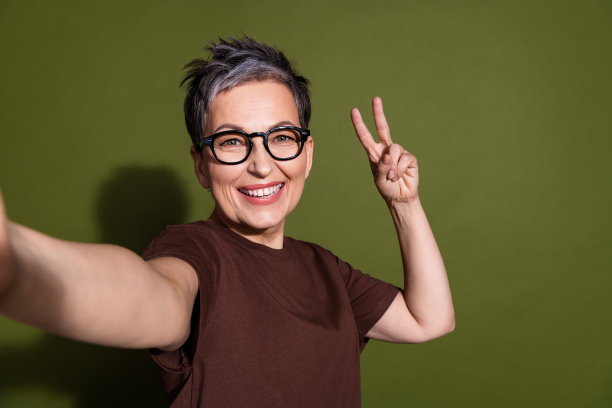 有魅力的退休妇女自拍V字手势，穿着时尚的棕色衣服，在卡其色背景下的照片人像图片下载