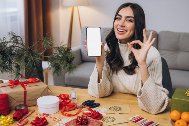 女人在包裹新年或圣诞礼物，同时拿着智能手机图片下载
